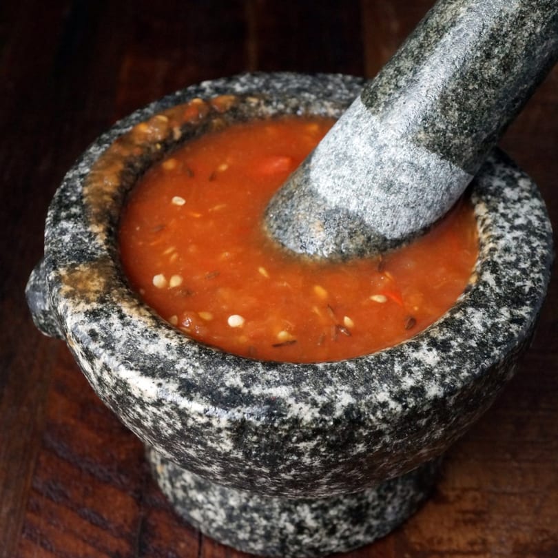 Close-up of a granite mortar and pestle with a vibrant orange Nepalese sauce inside.